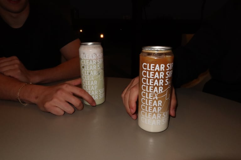 Two hands holding clear seltzer cans labeled CLEAR on a dark surface with ambient lighting