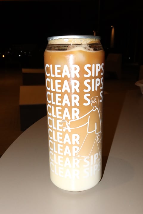 A tall beer can labeled Clear Sips with gradient tan to white coloring, filled with golden beer and ice, photographed against a dark background