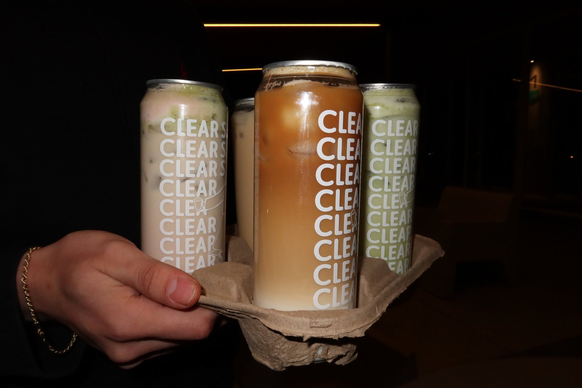Hand holding a cardboard six-pack carrier with three clear beer cans labeled CLEAR against a dark background